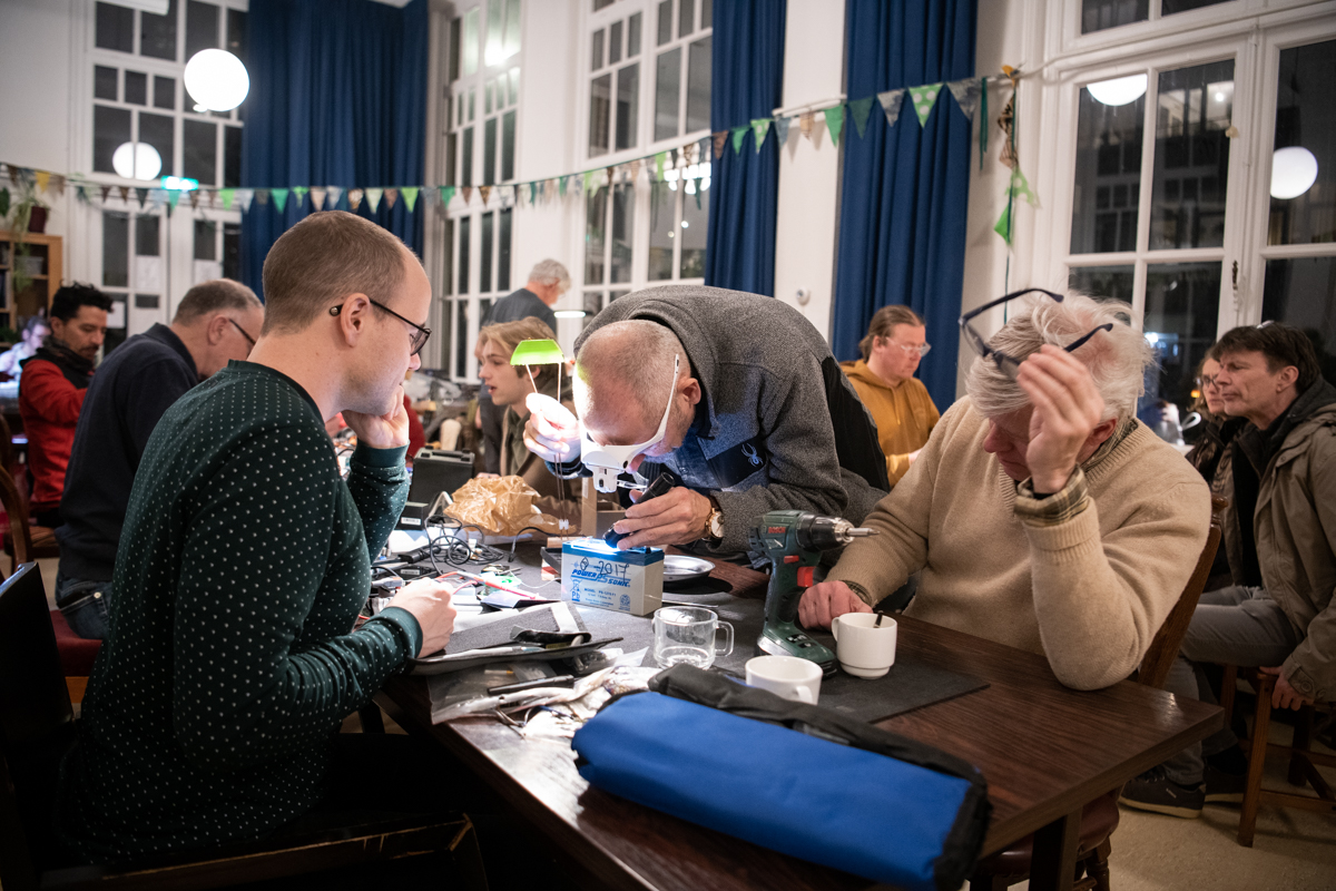 Avond bij Repair Café Jeltje: een reparatie aan een schroefmachine, met veel bezoekers en vrijwilligers.